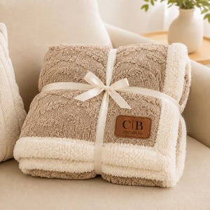 May include: A folded tan blanket with a textured cable knit pattern and a white sherpa fleece border. The blanket is tied with a white satin ribbon and features a brown leather patch with the embossed letters C|B and the date 2025.08.16.