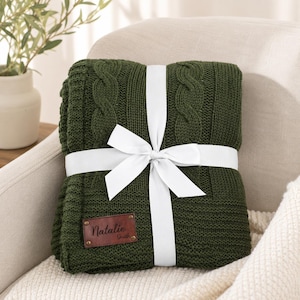 May include: A dark forest green knitted throw blanket featuring a cable knit pattern and a white ribbon tied in a bow around the center. A rectangular brown leather patch is attached to the lower corner with the name Natalie Smith embossed on it.