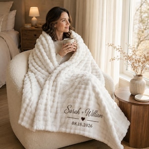 May include: A white textured throw blanket featuring the printed text Sarah + William and the date 08.18.2026 with a small heart icon. The blanket has a soft, raised grid pattern and is draped over a person sitting in a chair holding a mug.