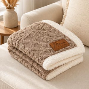 May include: A folded tan blanket with a textured cable knit pattern on one side and a white sherpa fleece lining on the other. A brown rectangular faux leather patch is stitched to the corner, featuring the text Mama Est. 2025 in black script.