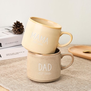 Puede incluir: Dos tazas de cerámica, una apilada encima de la otra. La taza superior es de color crema con la palabra "MAMA" y "EST 1985" en blanco. La taza inferior es de color beige con "DAD" y "EST 1998" en blanco. Ambas tazas tienen asas.