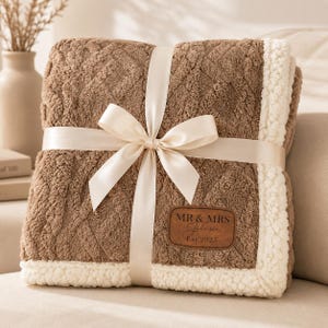 May include: A folded brown textured blanket with a white sherpa fleece border. The blanket features a diamond-patterned surface and a brown leather-like patch with the text MR & MRS Johnson Est. 2023. It is tied with a smooth cream-coloured ribbon.