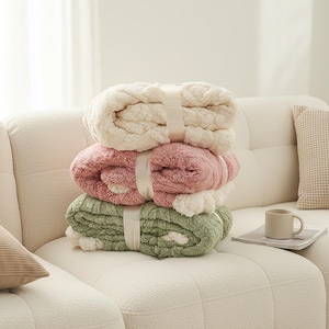 May include: A stack of three folded plush blankets secured with a white ribbon. The top blanket is cream, the middle is dusty rose, and the bottom is sage green. Each blanket features a textured, raised pattern and a soft, fuzzy fleece-like finish.