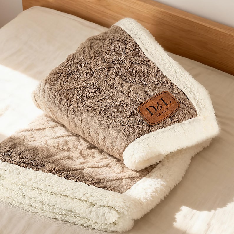 May include: A folded tan blanket with a textured diamond and cable knit pattern on one side and a white faux sheepskin lining on the other. A brown rectangular leather patch is sewn onto the corner, featuring the text D&L and 08.16.25.