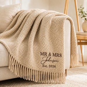 May include: A beige textured throw blanket with fringe trim, featuring dark brown embroidered text that reads MR & MRS Johnson Est. 2026. The blanket has a soft, nubby knit pattern and is draped over the arm of a light-coloured sofa.