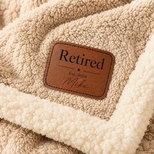 May include: A tan, textured fleece blanket with a white sherpa trim. A rectangular brown leather patch is stitched onto the center, featuring the black text "Retired", "Est. 2026", and the name "Mike" with a small star graphic in between.