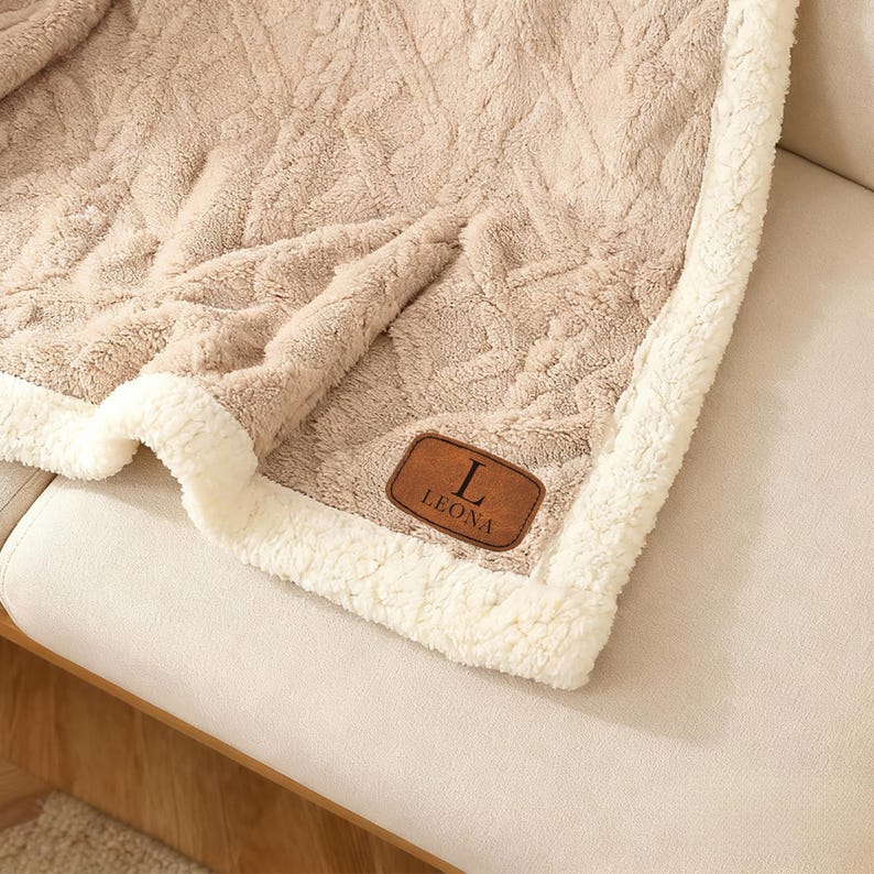 May include: A beige throw blanket with a textured cable knit pattern on one side and a soft white sherpa fleece lining. The blanket features a rectangular brown faux leather patch in the corner with the letter L and the name LEONA embossed in black.