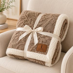 May include: A folded brown blanket with a textured leaf pattern and a white sherpa fleece border. The blanket is tied with a cream-coloured satin ribbon and features a brown leather-like patch with the letter L and the text LEONA embossed on it.