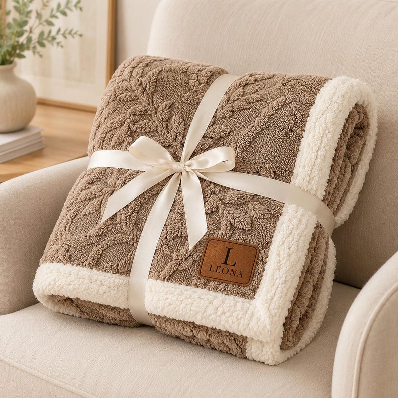 May include: A folded brown blanket with a textured leaf pattern and a white sherpa fleece border. The blanket is tied with a cream-coloured satin ribbon and features a brown leather-like patch with the letter L and the text LEONA embossed on it.