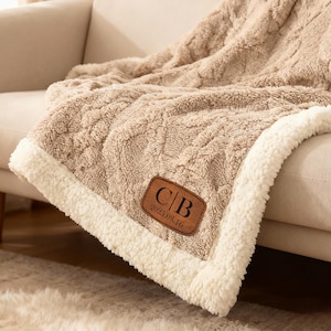 May include: A beige cable-knit throw blanket with a white sherpa fleece lining. The blanket features a brown rectangular leather patch with the letters C|B and the date 2025.08.16 embossed in black text on the front corner of the fabric.