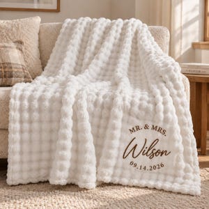 May include: A white textured throw blanket with a raised bubble pattern. The blanket features brown embroidered text that reads MR. & MRS. Wilson followed by the date 14.09.2026 in a script and serif font combination on the lower right corner.