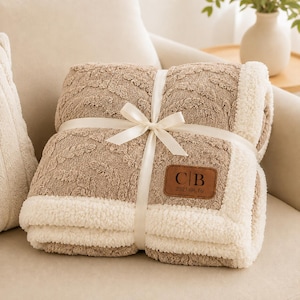 May include: A folded tan blanket with a textured cable knit pattern and a white sherpa fleece border. The blanket is tied with a white satin ribbon and features a brown leather patch with the embossed letters C|B and the date 2025.08.16.