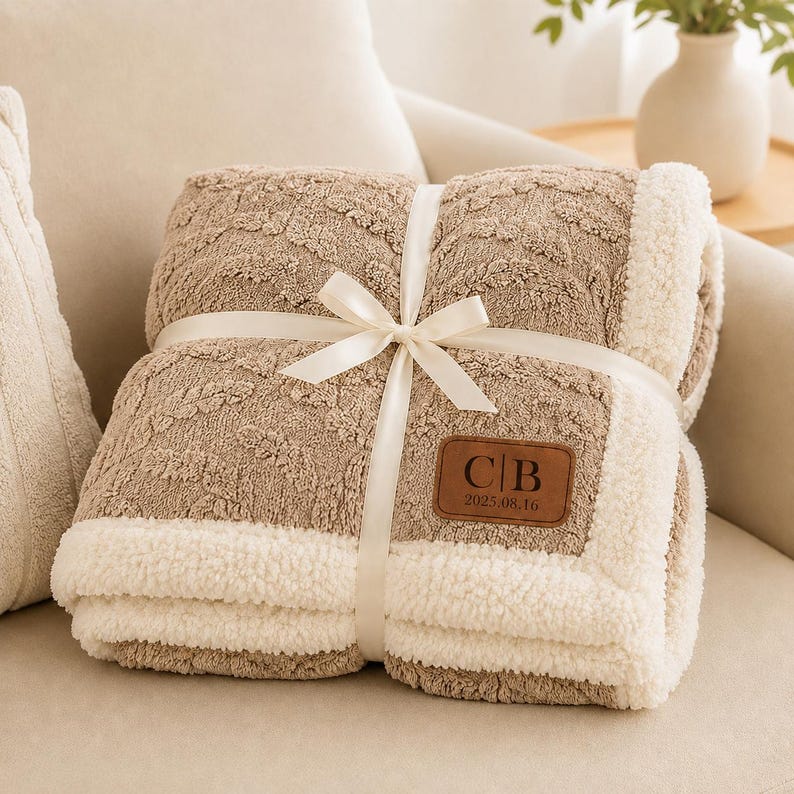 May include: A folded tan blanket with a textured cable knit pattern and a white sherpa fleece border. The blanket is tied with a white satin ribbon and features a brown leather patch with the embossed letters C|B and the date 2025.08.16.