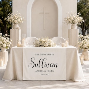 以下が含まれることがあります： 黒い文字で「THE NEWLYWEDS」「Sullivan」「AMELIA & HENRY」「10.03.2027」とプリントされた白いウェディングテーブルランナー。テーブルランナーは、白いフラワーアレンジメント、キャンドル、グラスで飾られたテーブルの上に掛けられています。