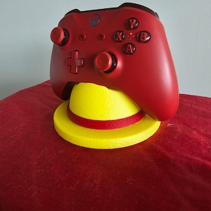 May include: A red Xbox controller sits on a yellow and red hat-shaped stand. The controller has black buttons and directional controls. The stand is yellow with a red band around the middle, set against a red background.