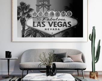 May include: Black and white framed print of the iconic "Welcome to Fabulous Las Vegas Nevada" sign. The sign is illuminated with lights and set against a backdrop of palm trees. The print hangs above a gray sofa.