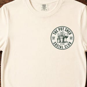 Puede incluir: Una camiseta de manga corta color crema con un gráfico circular negro en el pecho izquierdo. El diseño incluye una planta en maceta, una paleta de jardín y una copa de vino con el texto "THE POT SHED SOCIAL CLUB" y "EST. 2024" impreso.