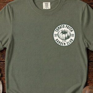 Può includere: Una t-shirt a maniche corte color verde oliva con una grafica circolare bianca sul petto sinistro. La grafica raffigura un pomodoro con il testo "TOMATO PATCH GARDEN CLUB" e "EST. 2026" insieme alle parole "GOOD DIRT" e "GOOD TASTE".