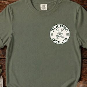 Könnte beinhalten: Ein olivgrünes Kurzarm-T-Shirt mit einer kreisförmigen Grafik auf der linken Brust. Die Grafik enthält den Text THE ALLOTMENT SOCIAL CLUB, EST. 2020, GOOD DIRT und GOOD VIBES sowie eine Abbildung einer gekreuzten Schaufel und Grabegabel.
