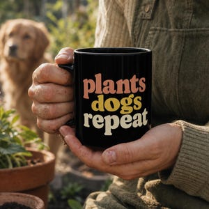 May include: A black ceramic mug featuring the text plants in orange, dogs in yellow, and repeat in cream. The mug has a cylindrical shape with a handle on the side. The text is printed in a bold, rounded, retro-style font on the front of the mug.