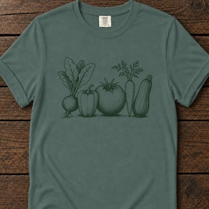 Könnte beinhalten: Ein salbeigrünes Kurzarm-T-Shirt mit einer dunkelgrünen Strichzeichnung einer Roten Bete, einer Paprika, einer Tomate, einer Karotte und einer Zucchini, die in einer Reihe in der Mitte angeordnet sind. Das T-Shirt hat einen Rundhalsausschnitt.
