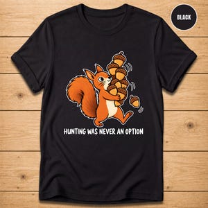 May include: A black t-shirt featuring a cartoon orange squirrel holding a large stack of brown acorns. Below the graphic, the white text reads: HUNTING WAS NEVER AN OPTION. A small circular badge in the top right corner displays the word BLACK.