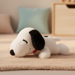 Puede incluir: Un peluche de Snoopy en una pose relajada, tumbado sobre una superficie beige. El perro blanco tiene orejas negras y una mancha negra en la espalda, con un collar rojo. El juguete es suave y tierno.