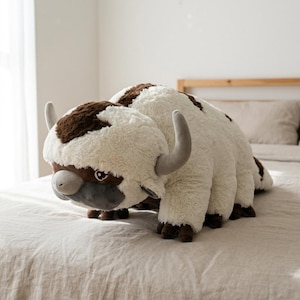 May include: A plush toy shaped like a bison with a white body, brown patches, and gray horns. The toy has a gray snout and brown hooves. It is lying on a bed with a neutral-colored bedspread.