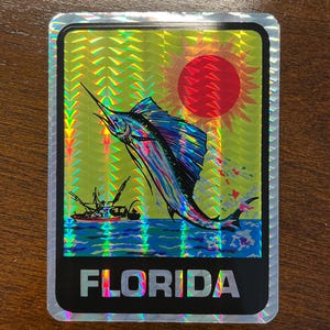 May include: A rectangular holographic sticker featuring a vibrant illustration of a sailfish leaping from blue water against a yellow background with a red sun. A small fishing boat is visible in the distance. The word FLORIDA is printed in white.
