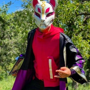Peut inclure: Un masque de renard blanc et rouge avec des détails dorés, porté avec un sweat à capuche rouge et une veste noire avec une doublure violette. Le costume est un cosplay, probablement pour un personnage de jeu vidéo.
