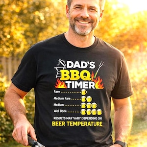May include: Black t-shirt with the text "DAD'S BBQ TIMER" and a beer-themed cooking guide. The shirt features a grilling fork and flames design. The wearer is grilling food outdoors.
