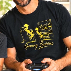 May include: A black t-shirt featuring a yellow graphic of two individuals sitting in gaming chairs while playing a racing video game on a television screen. The text "Gaming Buddies" is printed in a yellow script font below the illustration.
