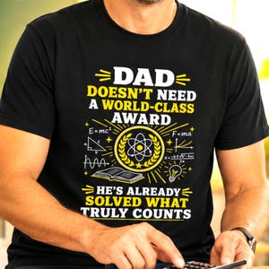 May include: A black short-sleeved t-shirt featuring yellow and white graphic text that reads DAD DOESN'T NEED A WORLD-CLASS AWARD HE'S ALREADY SOLVED WHAT TRULY COUNTS, accompanied by scientific symbols including an atom, a book, and equations.
