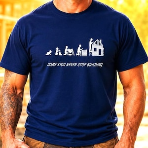 May include: A navy blue short-sleeved t-shirt featuring a white graphic print that depicts the evolution of a builder from a crawling infant to an adult constructing a house. The text "SOME KIDS NEVER STOP BUILDING" is printed in white below the graphic.
