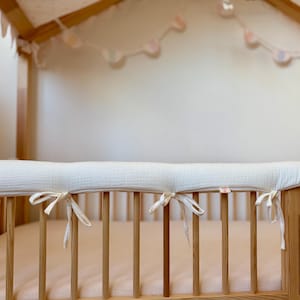 May include: A light cream-colored fabric crib bumper pad with tie-fasteners attached to the wooden slats of a crib. The crib features a light wood frame, a soft pink mattress, and a canopy top with hanging decorative elements and bunting.