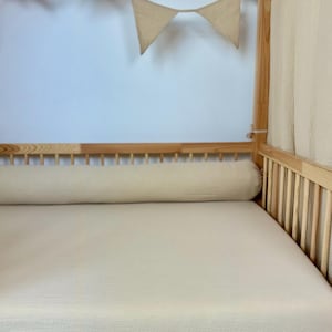Peut inclure: Un traversin en mousseline beige clair posé sur un drap-housse assorti dans un cadre de lit en bois naturel en forme de maison. Le lit est décoré d'une guirlande lumineuse en forme d'étoiles et de fanions triangulaires en tissu suspendus.