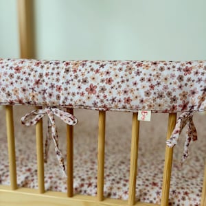 Könnte beinhalten: Ein gepolsterter Bettgitter-Schutz aus weißem Stoff mit einem wiederkehrenden Muster aus kleinen braunen, rosafarbenen und beigen Blumen. Der Schutz ist mit Stoffbändern an den Holzstäben befestigt und hat ein kleines Etikett mit der Aufschrift Esty.