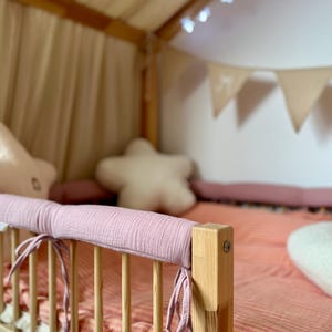 Puede incluir: Cama de madera con colchón a rayas rosas y protector a juego atado a la barandilla. Cojines con forma de estrella y nube decoran la cama. Se ve un dosel y luces de cadena.