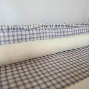 May include: A long, cylindrical bolster pillow in a solid cream colour, positioned in front of a rectangular crib bumper pad. The bumper features a blue and tan checkered pattern with a ruffled top edge, creating a coordinated bedding set.