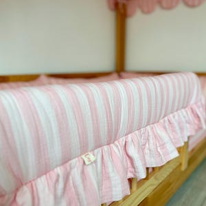 May include: A crib bumper pad featuring a light pink and white vertical striped pattern made of a textured fabric. The bumper includes a ruffled trim along the bottom edge and a small white fabric tag with text attached to the side of the ruffle.