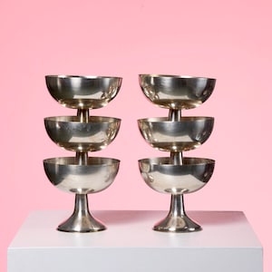Peut inclure: Six coupes à champagne argentées, empilées par paires. Chaque verre présente une coupe arrondie et un pied. L'ensemble est disposé sur une surface blanche devant un fond rose, créant une présentation élégante et festive.