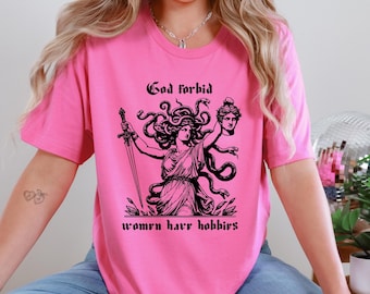 Puede incluir: Camiseta rosa con un gráfico negro de Medusa sosteniendo una espada y una cabeza cortada, con el texto "God forbid women have hobbies". El diseño está centrado en la camiseta.