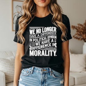 Op de afbeelding: Zwart T-shirt met witte tekst. De tekst luidt: "WE HEBBEN GEEN VERSCHIL MEER IN POLITIEKE MENING WE HEBBEN EEN VERSCHIL IN MORAAL." Het T-shirt wordt gedragen door een persoon in een spijkerbroek.