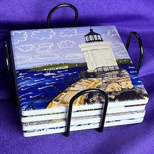 May include: A set of four square coasters with a lighthouse scene, held in a black metal rack. The coasters feature a white lighthouse on a rocky shore, blue water, and a cloudy sky. The rack has a curved handle.