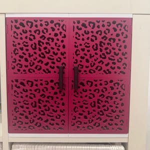 Può includere: Un mobile con due ante magenta con un motivo leopardato nero. Le ante hanno maniglie marrone scuro e sono inserite in una cornice bianca. Il mobile fa parte di un'unità di stoccaggio più grande.