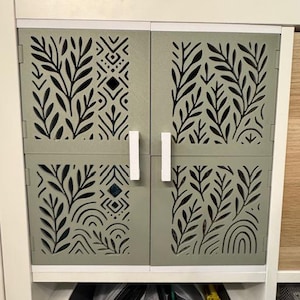 May include: A cabinet with sage green doors featuring laser-cut leaf and geometric designs. The doors have white handles and are mounted on a white frame. The cabinet is part of a larger storage unit.