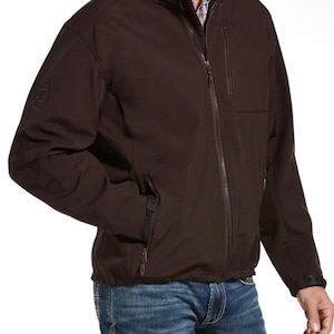 Puede incluir: Chaqueta marrón oscuro con cremallera, cuello alto y múltiples bolsillos. La chaqueta tiene un logotipo sutil en la manga izquierda y el pecho. La persona lleva vaqueros azules. La chaqueta está diseñada para actividades al aire libre.