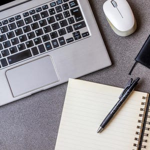 Pode incluir: Vista aérea de um espaço de trabalho com um laptop prateado, um mouse branco, um caderno preto e um bloco de notas em espiral. Uma caneta preta está sobre o bloco de notas. Os itens estão dispostos em uma superfície cinza.