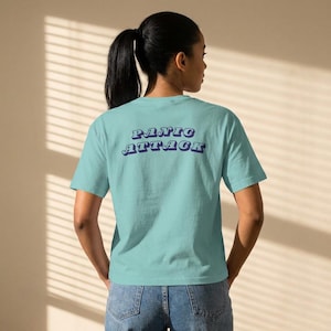Könnte beinhalten: Ein hellblaues T-Shirt mit dem Schriftzug "PANIC ATTACK" in einem Retro-Schriftzug auf dem Rücken. Das kurzärmelige Shirt scheint aus einem weichen Material zu sein. Das Model trägt eine blaue Jeans.