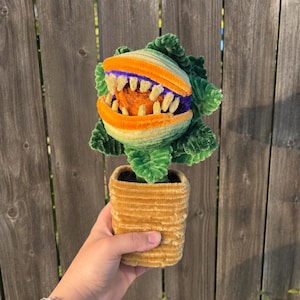 May include: A plush toy shaped like a carnivorous plant with a wide-open mouth, sharp teeth, and a bright orange centre. The plant has textured green leaves and is potted in a golden-yellow, ribbed, rectangular fabric container.
